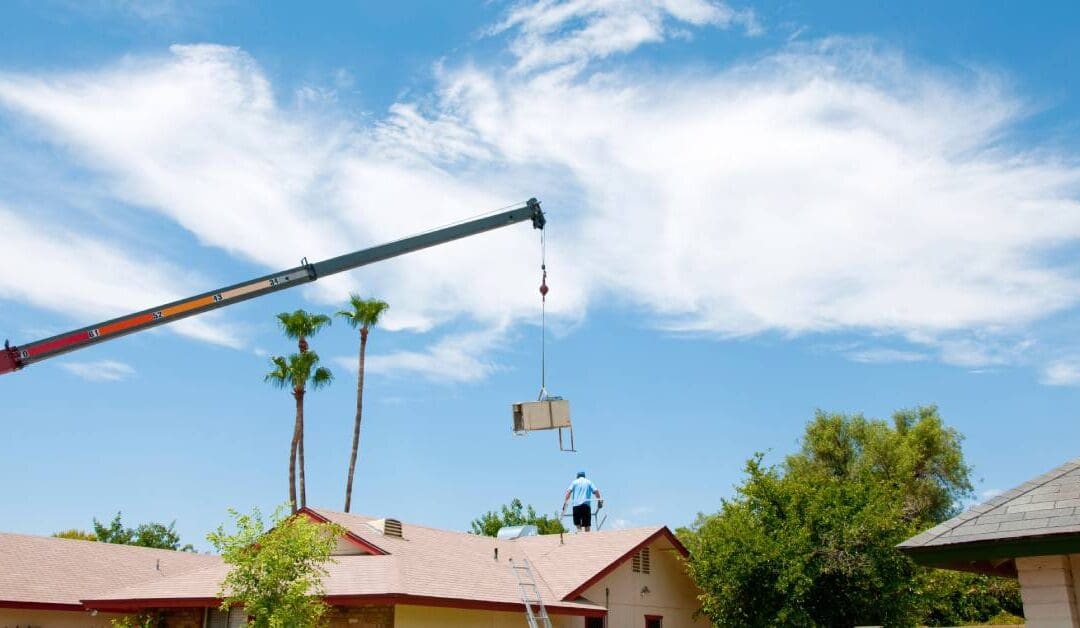 A crane holds an air conditioning unit in the sky over a group of homes. There are trees and a big cloud in the background.