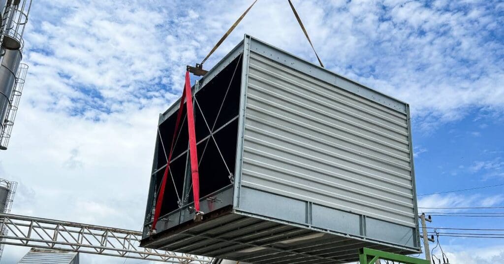 A crane holds a cooling tower with a series of ropes. Other meal architecture and power lines are in the background.