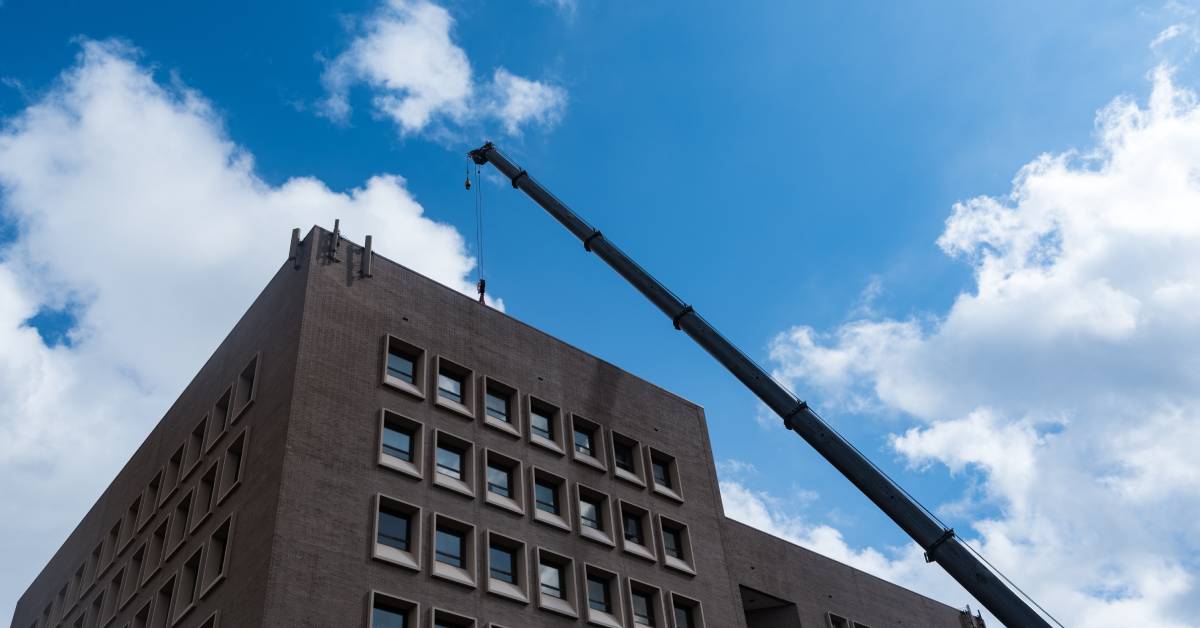 A large crane hoists an HVAC system to the top of a really tall and dark building with a lot of windows.