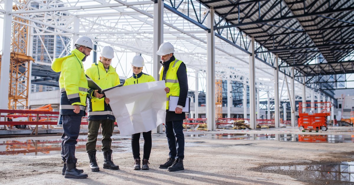 Engineers stand at a worksite with large yellow jackets and construction helmets, looking over a blueprint.