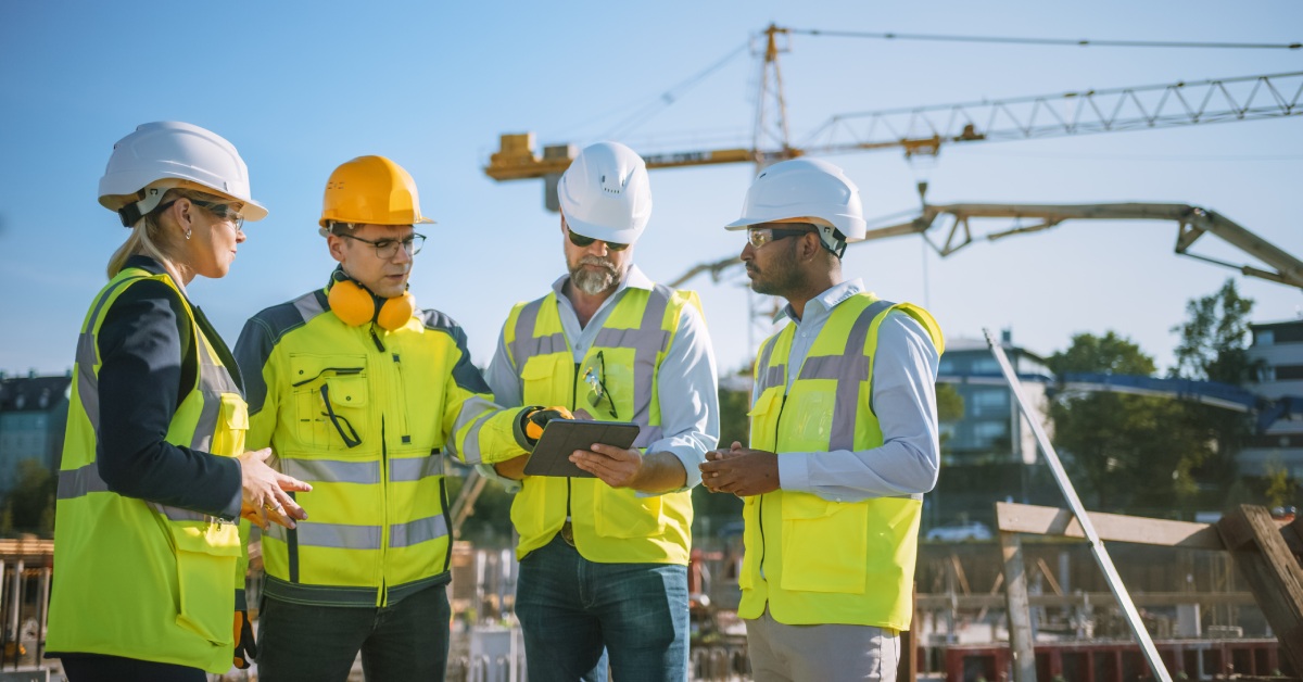 Some construction workers and engineers at a worksite with a crane in the background look at a tablet.