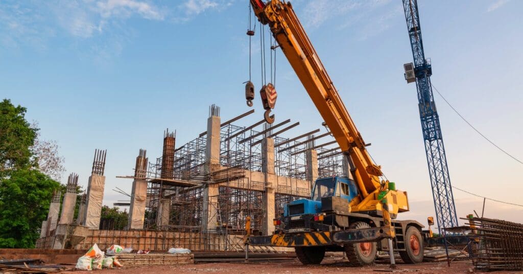 A mobile crane sits outside of a construction site. The ground is just dirt and there are trees in the background.