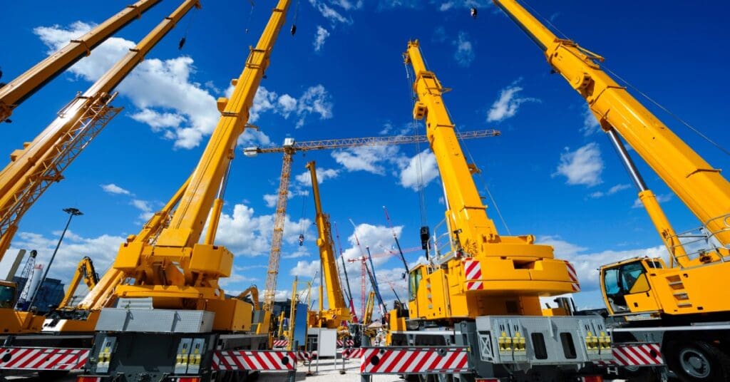 A lot sits full of different yellow construction cranes, all pointing towards the big blue sky and clouds.