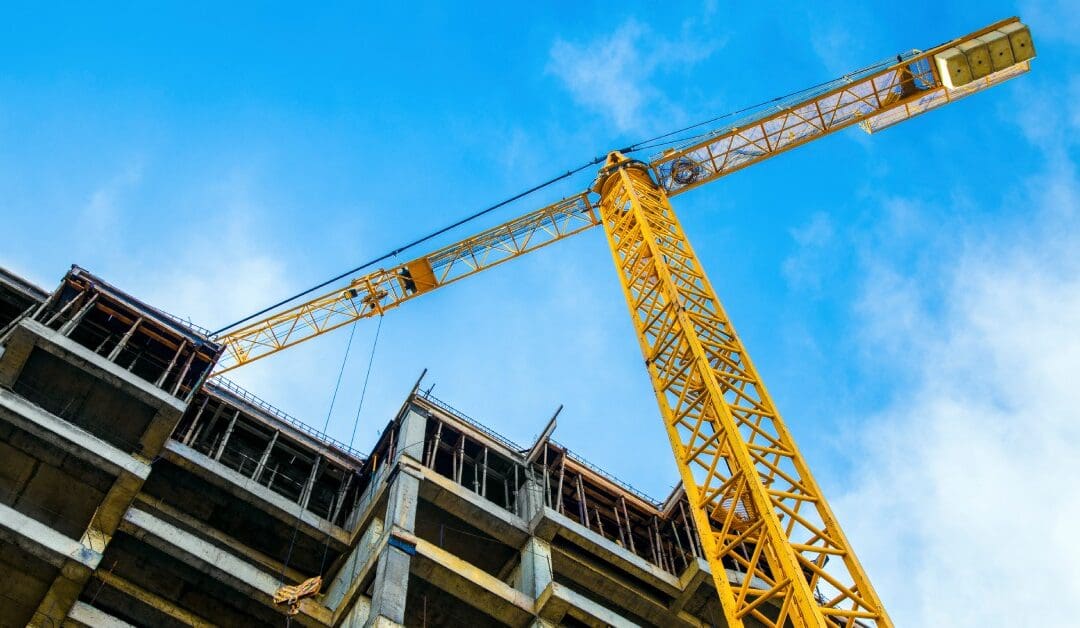 A yellow crane stands next to a building mid-construction with exposed beams and concrete. The sky is bright blue.