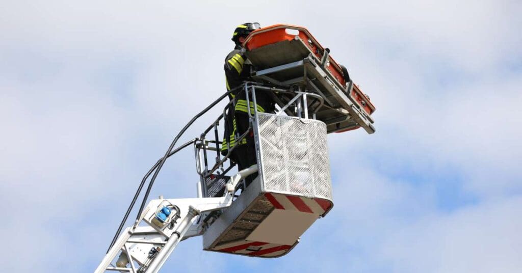 A firefighter stands on an aerial platform at the top of a crane, wearing standard firefighter gear and holding a stretcher.