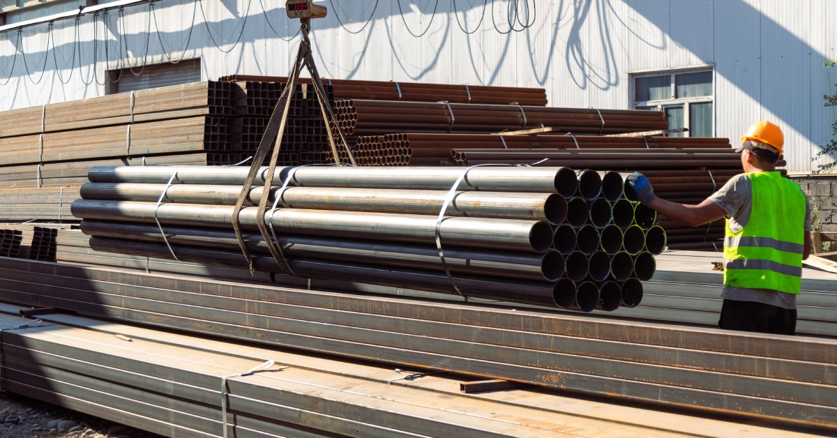 A worker in a hard hat guides a crane in picking up a couple dozen long metal pipes next to other building materials.