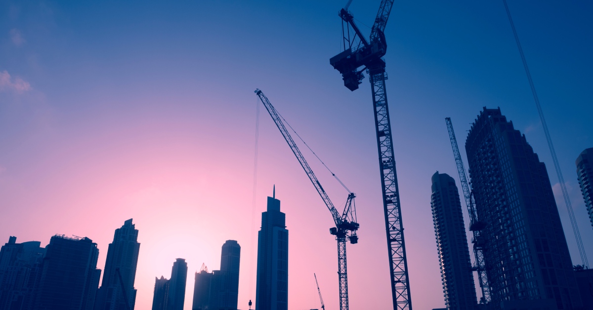 A skyline at sunset with numerous tall cranes standing tall next to numerous skyscrapers and unfinished buildings.