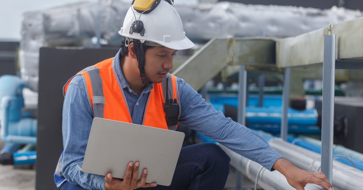 A man in a bright orange vest, white hard hat, and yellow earmuffs holds a laptop while inspecting the pipes of an HVAC unit.