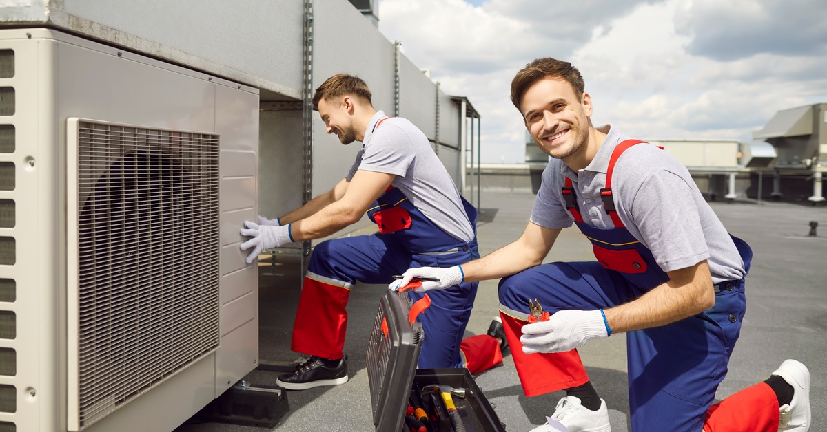 Two men dressed in similar orange-and-blue overalls smiling as they use tools to work on an HVAC unit.