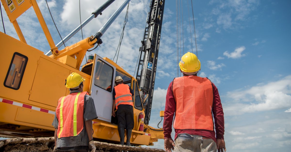 Two people are looking at a crane truck, while a third person climbs into the vehicle. Everyone is wearing safety gear.