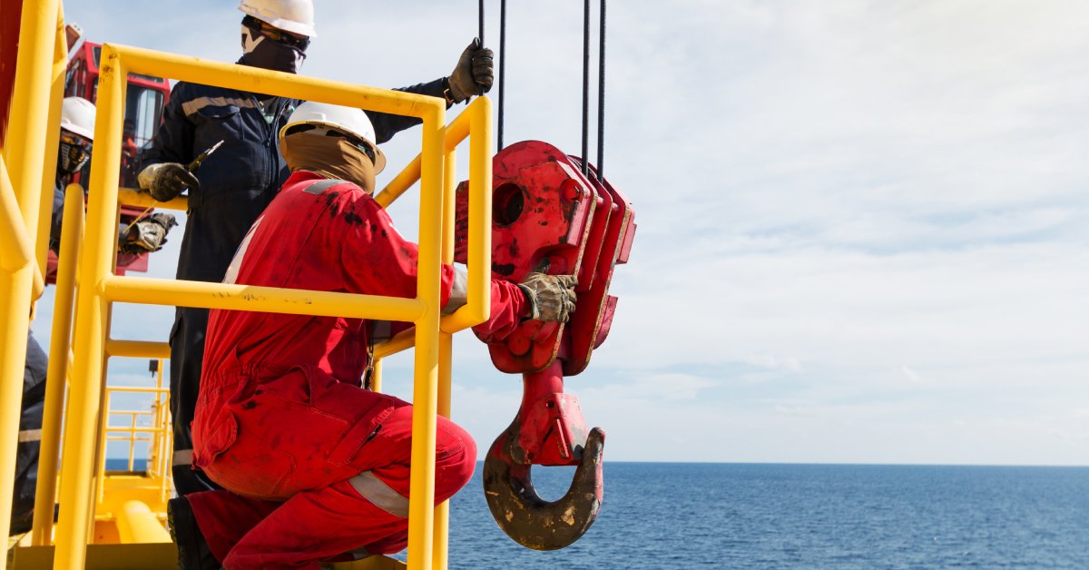 Two people wearing safety equipment inspect a large piece of rigging equipment while over a body of water.
