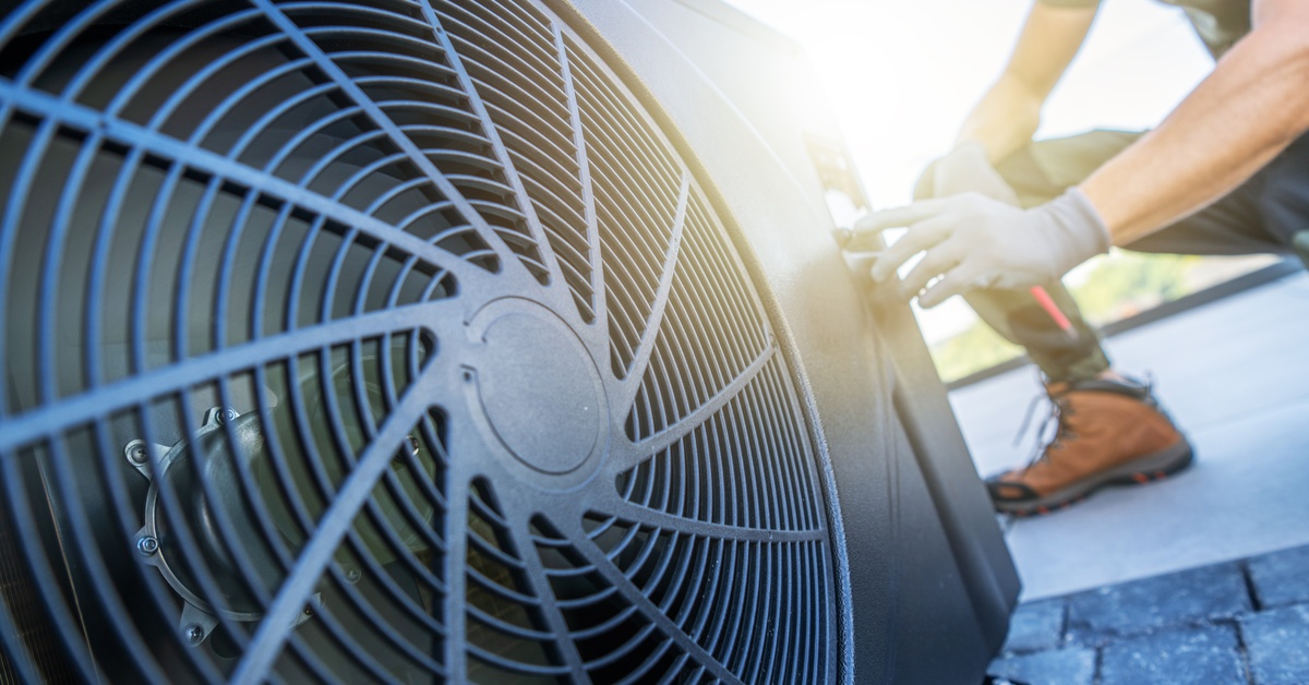 A technician crouching down next to an HVAC unit on a rooftop. The sun is shining between the person and the machine.