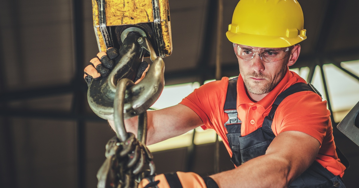 In a warehouse, a man wearing work gloves and a hard hat attaches rigging equipment to the hook of a crane.
