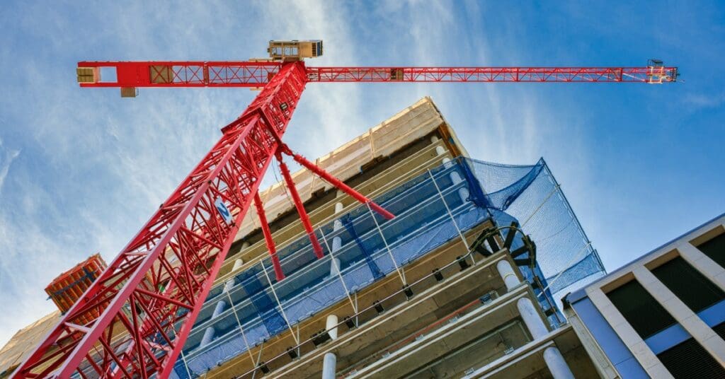 From ground level looking up, there's a partially constructed high-rise structure with a red crane next to the work site.