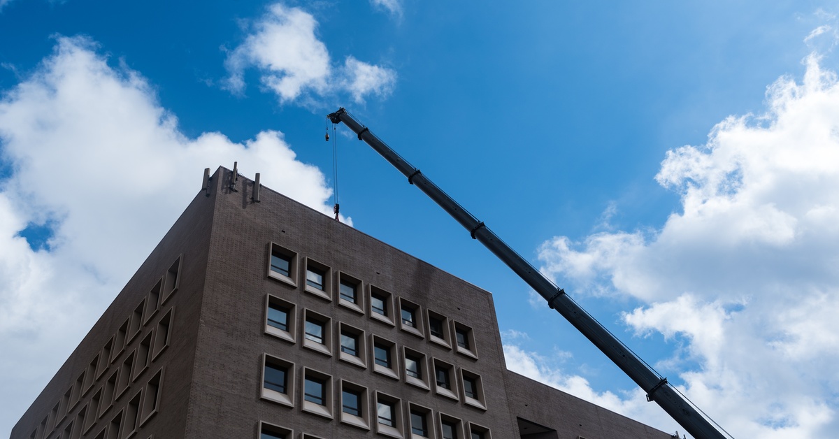 An HVAC system being lifted by a crane onto a building. Behind the building and crane, the sky is bright with white clouds.