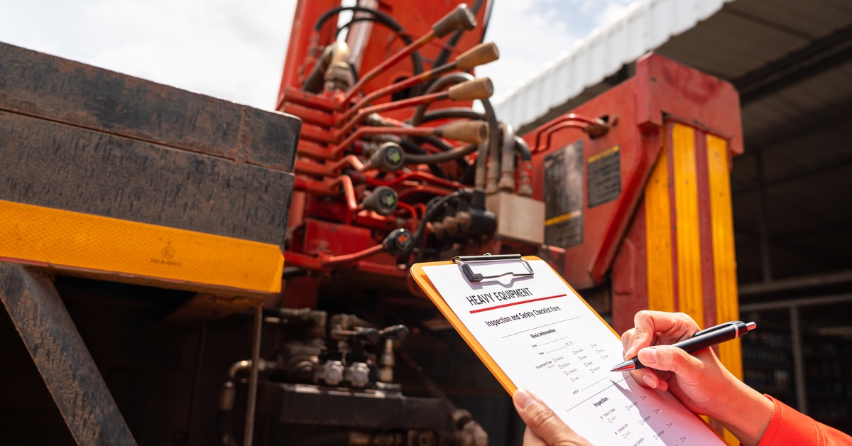 A person holding a clipboard and pen while inspecting a mobile crane vehicle. The crane is blurred behind the clipboard.