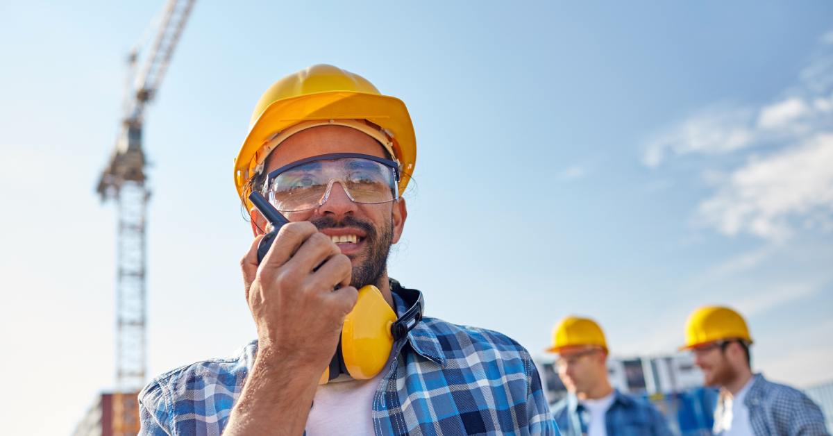 A crew member with a hard hat on holding a walkie talkie up to speak into it. Behind him is a tower crane.