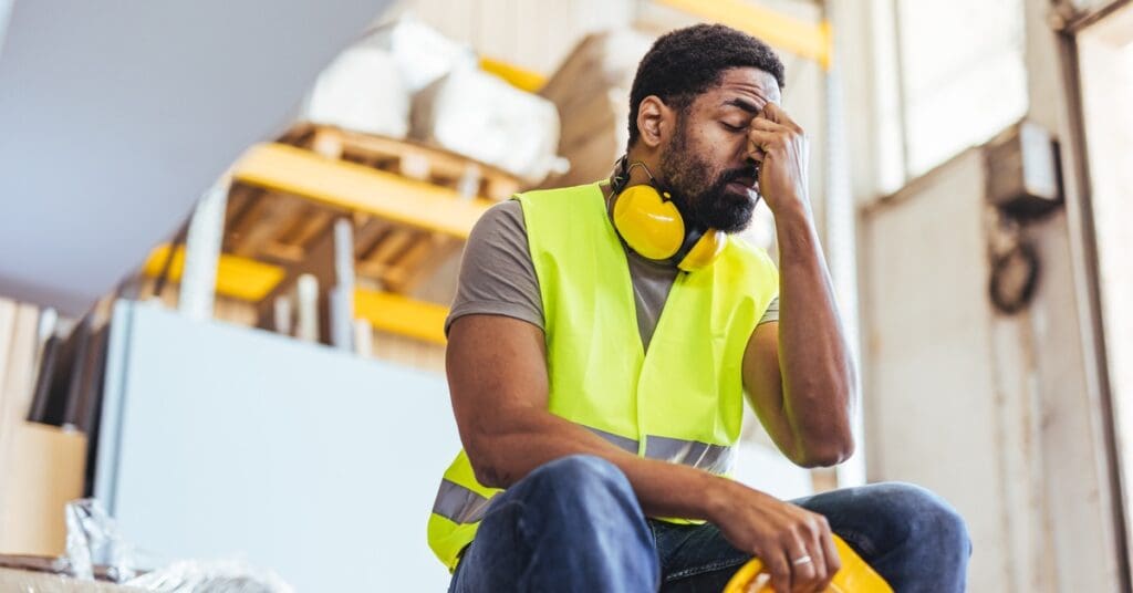 A weary construction worker in safety gear sitting down at a work site, resting his face in his hand.