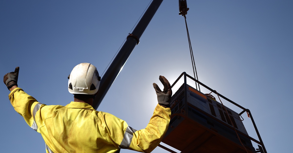 A rigger wearing dirty safety gear directs a crane with hand signals on where to place a piece of equipment.