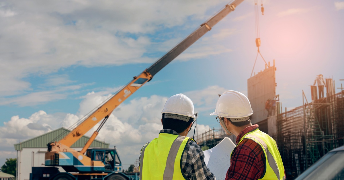 Two engineers wearing hard hats discuss plans for a crane lift. In front of them, the crane is lifting concrete.