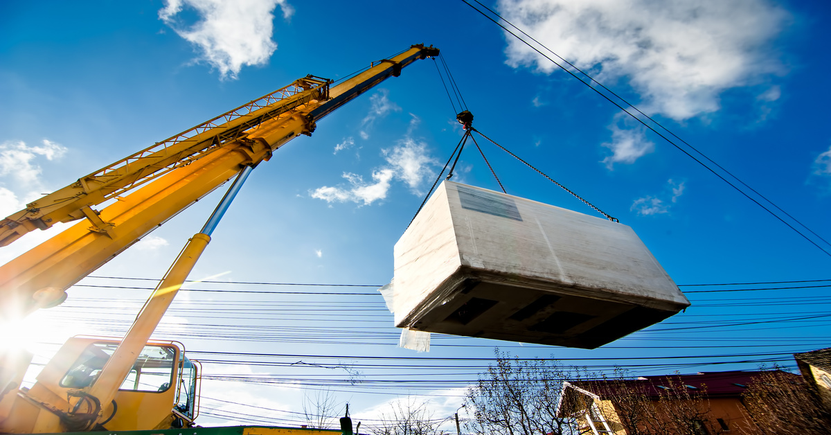 A crane lifting an electric generator into the air with rigging and a hook around it on a sunny day.