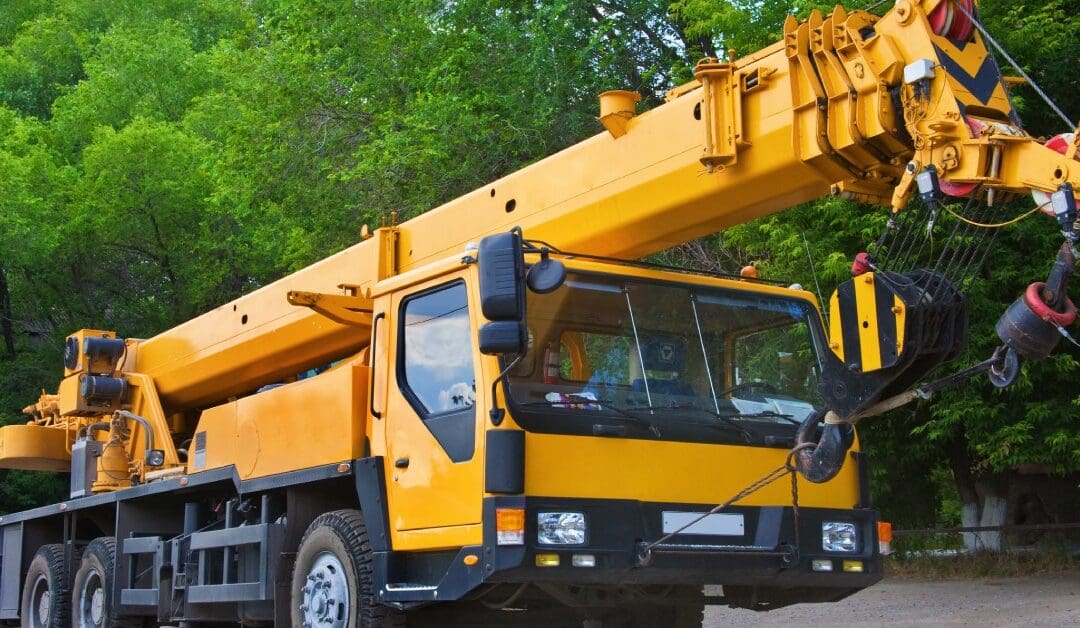 A powerful mobile crane unit parked on a road with the crane retracted into the arm. Behind the crane is a forest area.
