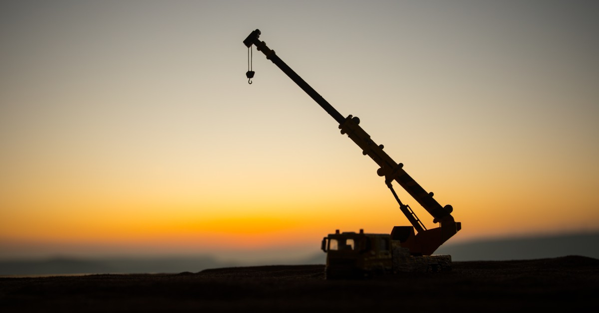 A mobile crane unit with the crane extended upward into the sky. The crane is a shadow illuminated by the sunset.