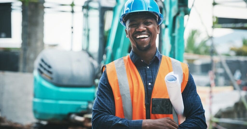 A young man on a construction site wearing safety gear with a blueprint under his arm. There is a crane behind him.