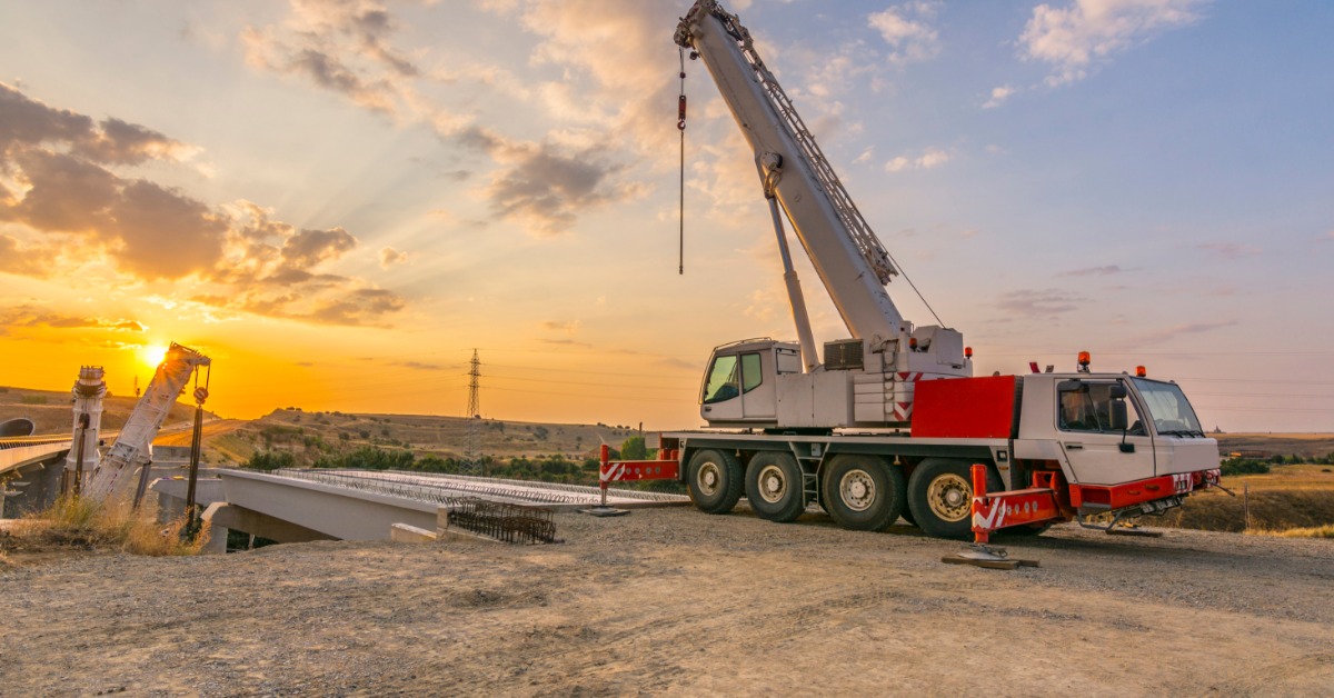 A mobile construction crane on a work site, helping to build a bridge. The sun is setting in the distance.