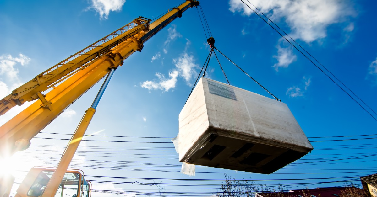 An industrial crane lifting a wrapped electric generator into the air. The sky behind the generator is clear.